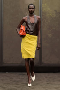Model walking the runway in a brown sleeveless top, bright yellow pencil skirt, white heels, and an orange clutch.