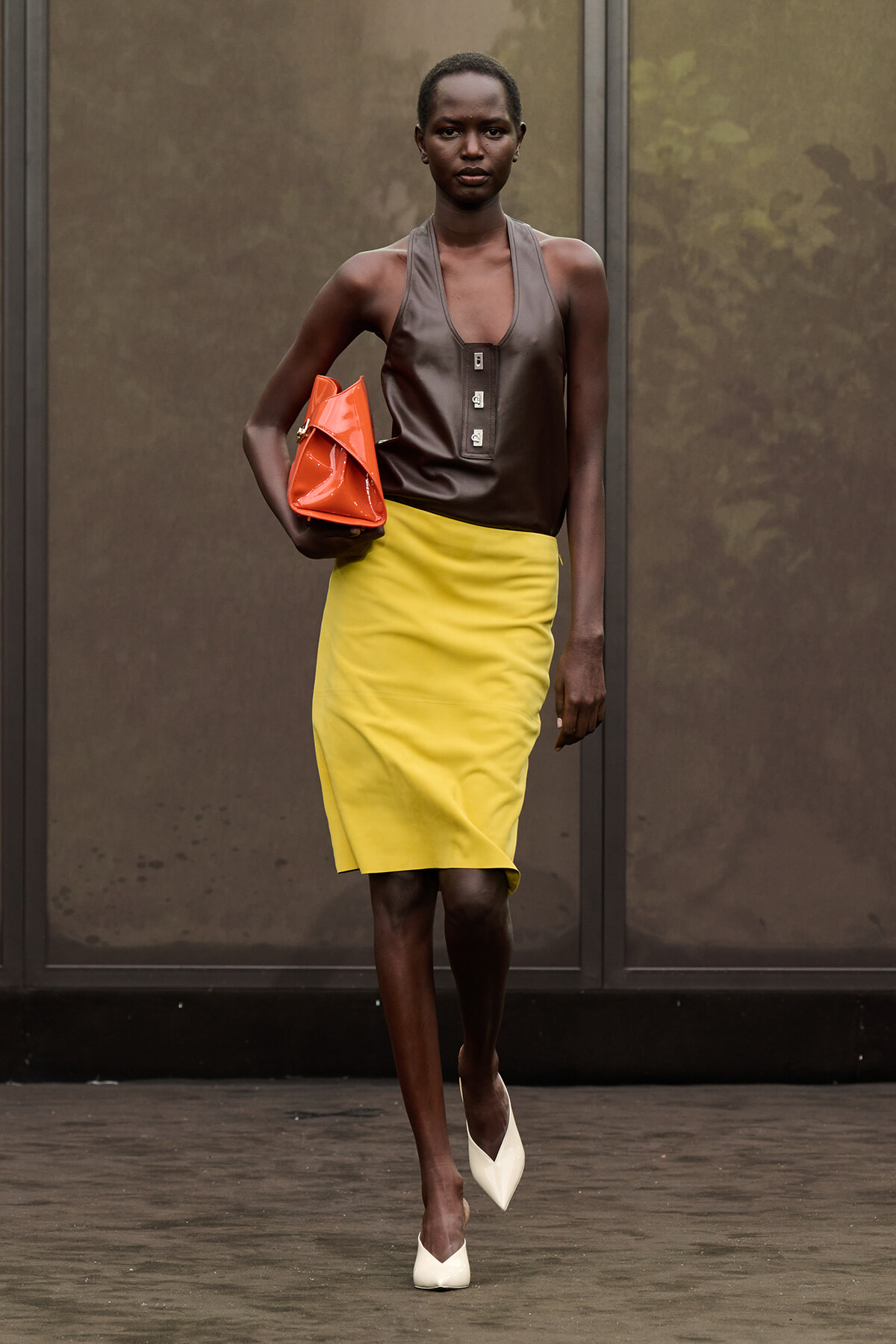 Model walking the runway in a brown sleeveless top, bright yellow pencil skirt, white heels, and an orange clutch.