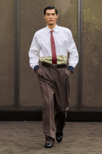 Man in a white dress shirt, maroon tie, and brown pleated trousers walks toward the camera with hands in pockets outside a building.