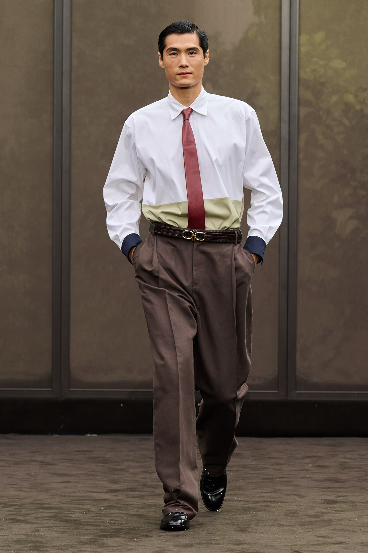 Man in a white dress shirt, maroon tie, and brown pleated trousers walks toward the camera with hands in pockets outside a building.