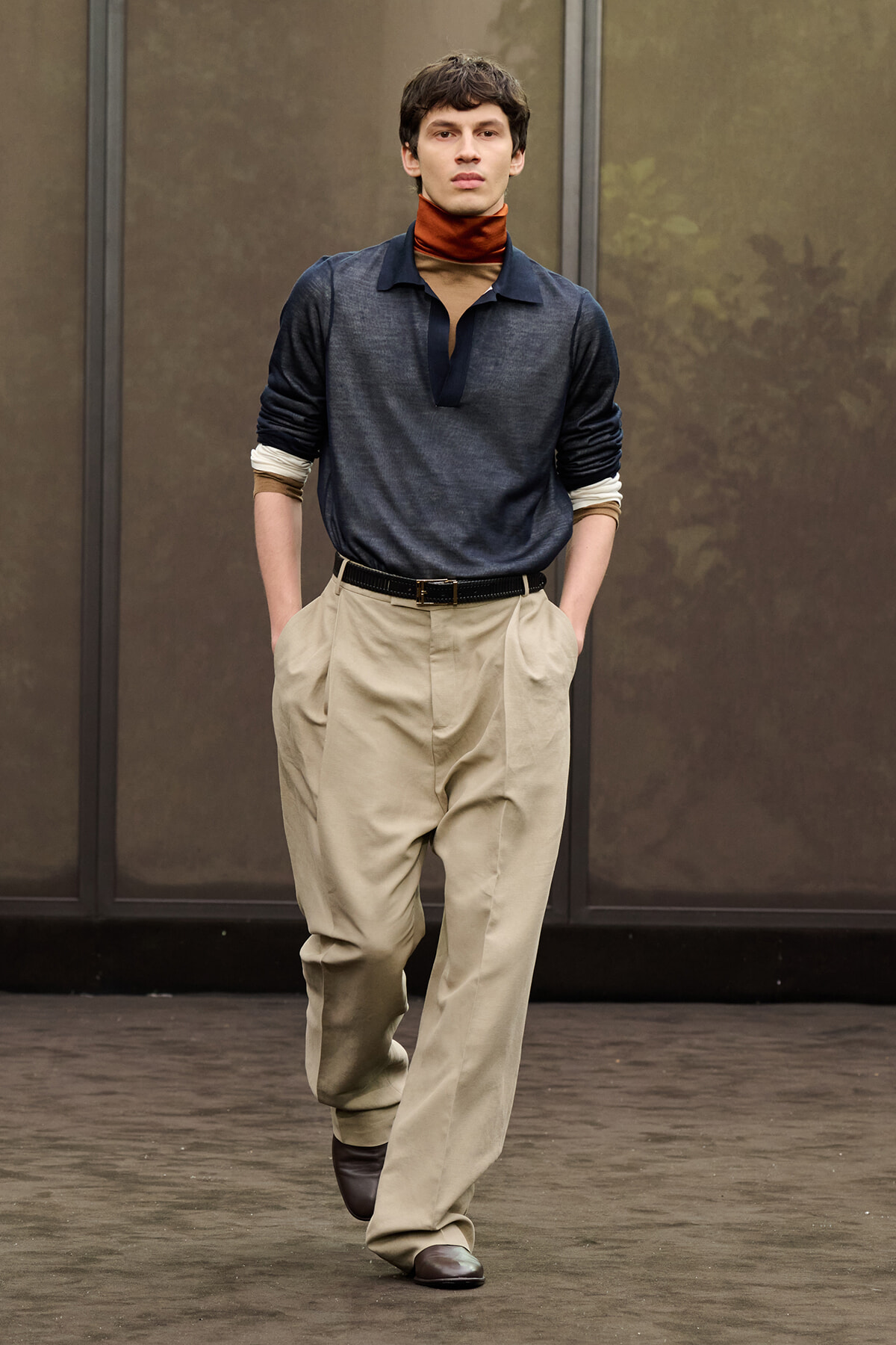 Male model walking the runway in a navy polo with a brown scarf, beige wide-leg trousers, a black belt and brown shoes.