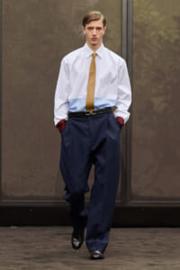 Male model walking toward camera in a white shirt with light blue yoke, mustard tie, navy wide trousers, and burgundy cuff accents on a runway.