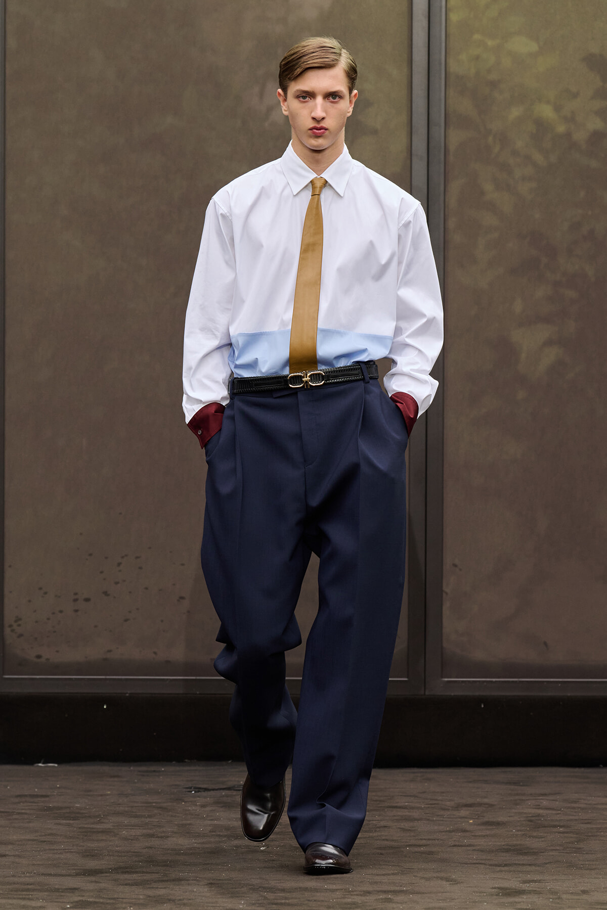 Male model walking toward camera in a white shirt with light blue yoke, mustard tie, navy wide trousers, and burgundy cuff accents on a runway.