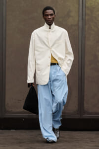 Male model in an oversized cream blazer over a mustard top, tucked into light blue baggy trousers, carrying a black bag.