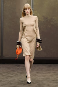 Model walking a runway in a beige, knee-length dress with fringe panels at the hips and black-heeled shoes. She carries an orange clutch.