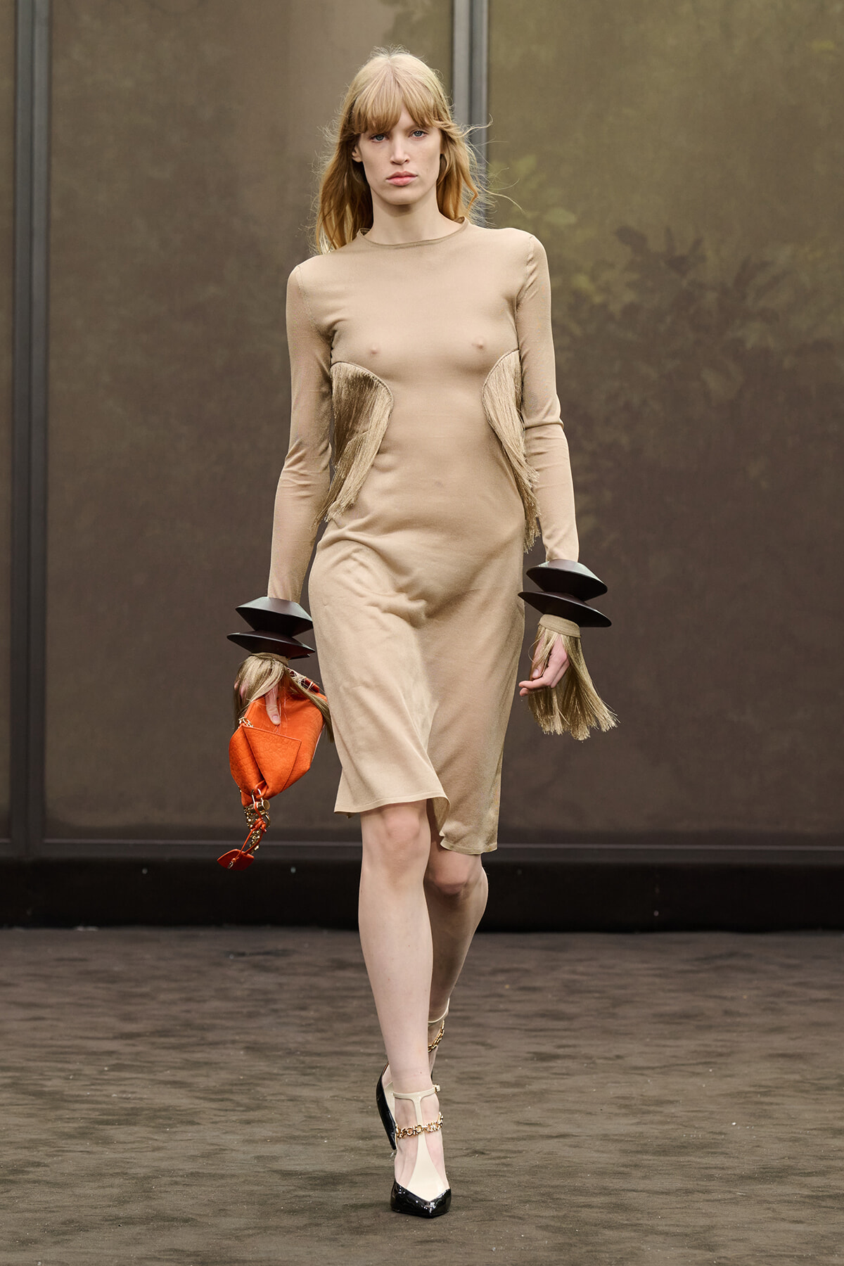 Model walking a runway in a beige, knee-length dress with fringe panels at the hips and black-heeled shoes. She carries an orange clutch.