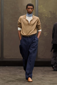 Full-body runway shot of a male model wearing a tan top with layered turtlenecks, navy wide-leg trousers, a brown belt, and tan shoes, hands in pockets.