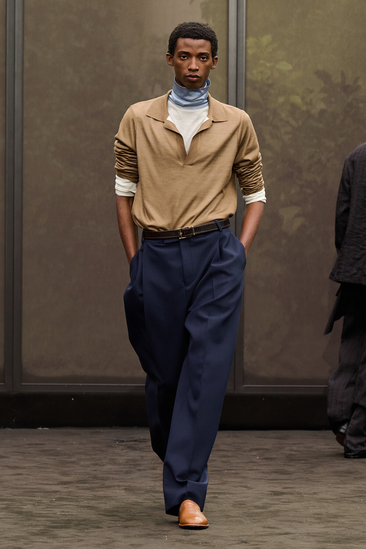 Full-body runway shot of a male model wearing a tan top with layered turtlenecks, navy wide-leg trousers, a brown belt, and tan shoes, hands in pockets.
