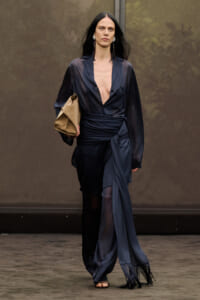 Runway model in a navy sheer wrap blouse with deep V, matching pants, and a tan clutch.