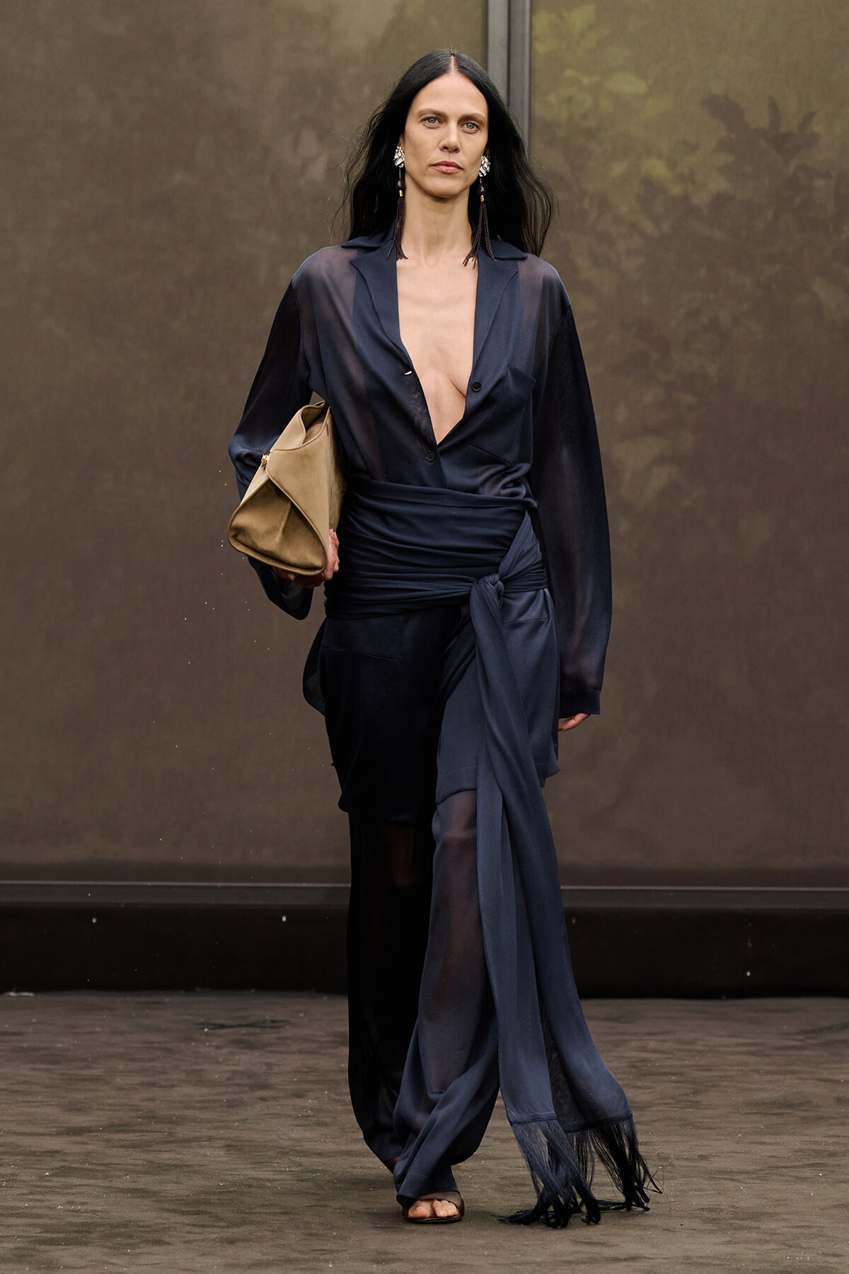 Runway model in a navy sheer wrap blouse with deep V, matching pants, and a tan clutch.