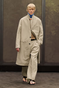 Model walking on a runway in a long beige overcoat over matching suit pieces, with a blue mock neck underneath. The look is minimalist and monochrome.