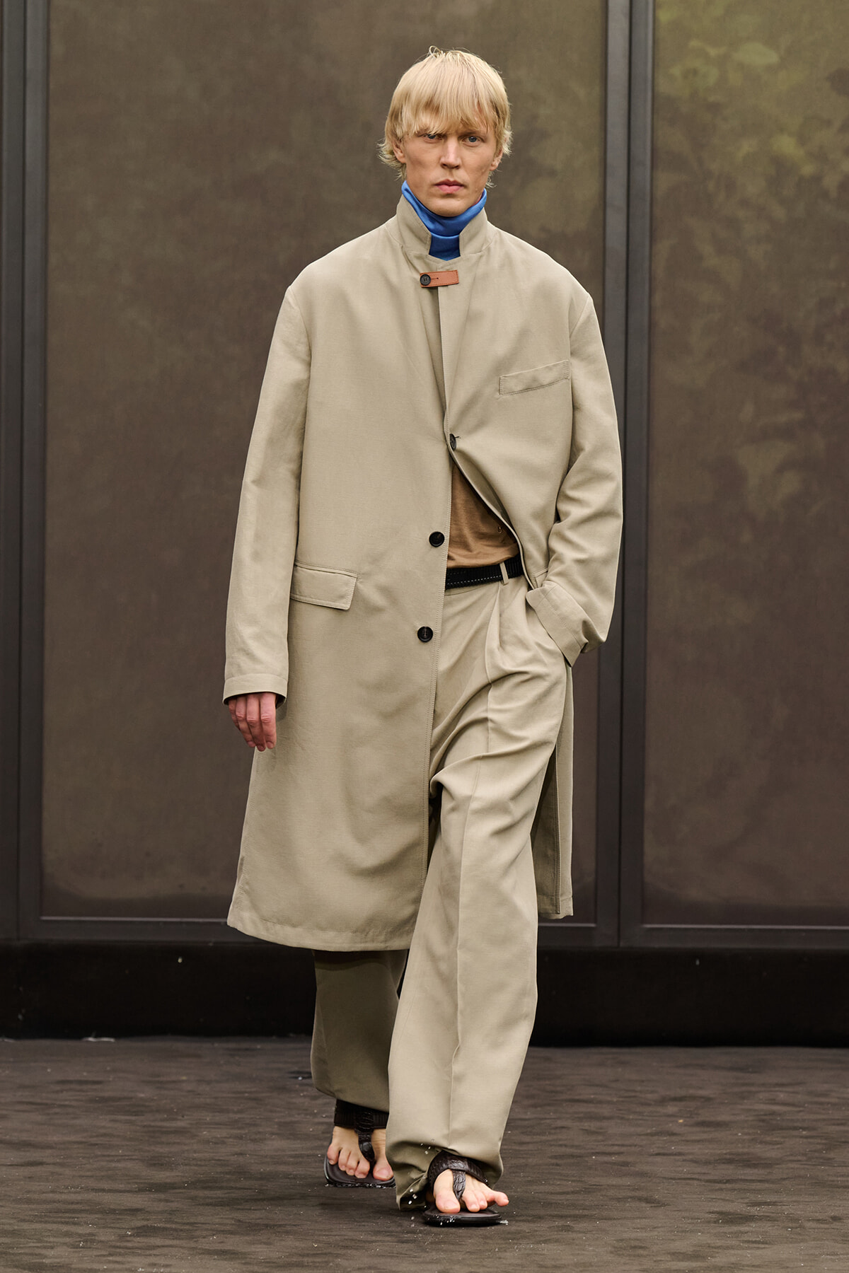 Model walking on a runway in a long beige overcoat over matching suit pieces, with a blue mock neck underneath. The look is minimalist and monochrome.