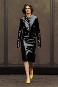 Model on a runway wearing a shiny black patent leather cropped jacket and matching knee-length skirt, with yellow pointed-toe heels.