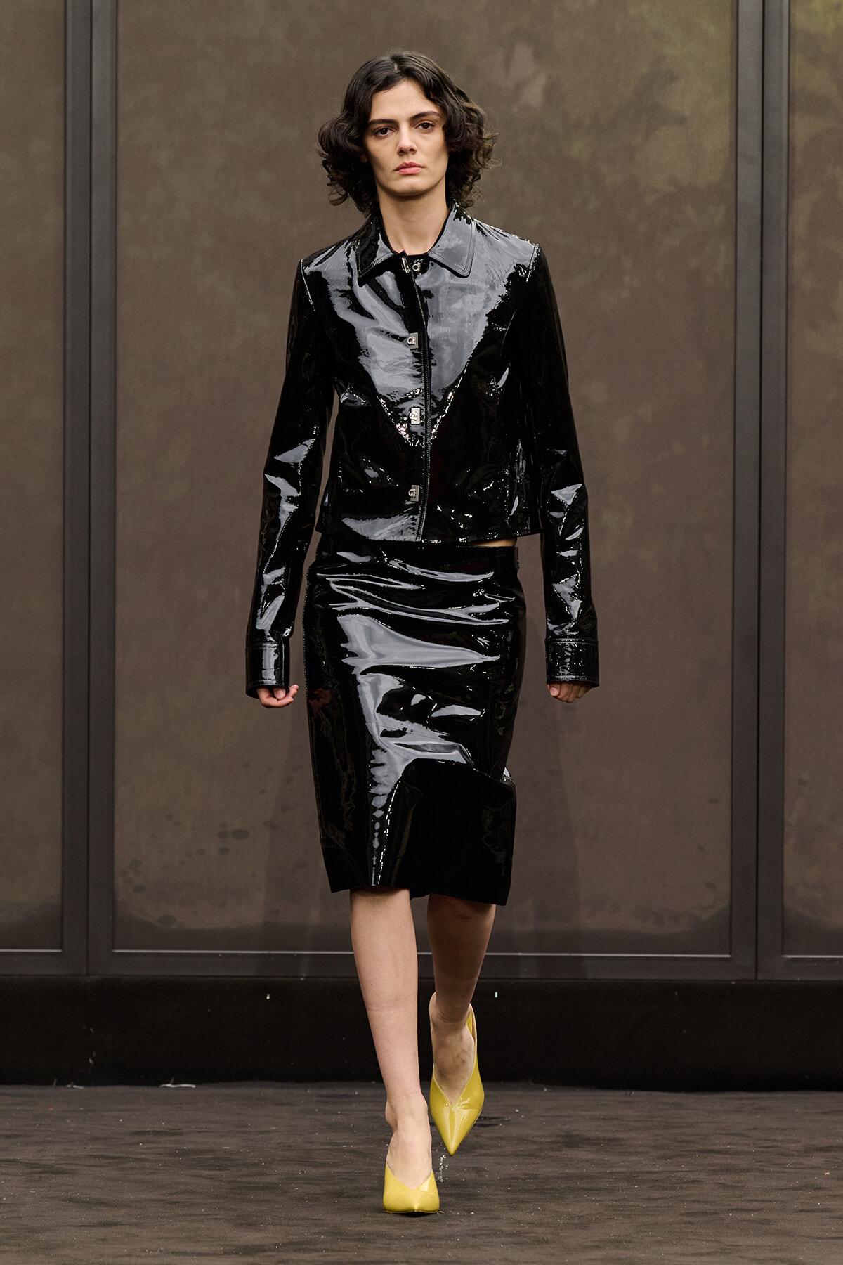 Model on a runway wearing a shiny black patent leather cropped jacket and matching knee-length skirt, with yellow pointed-toe heels.