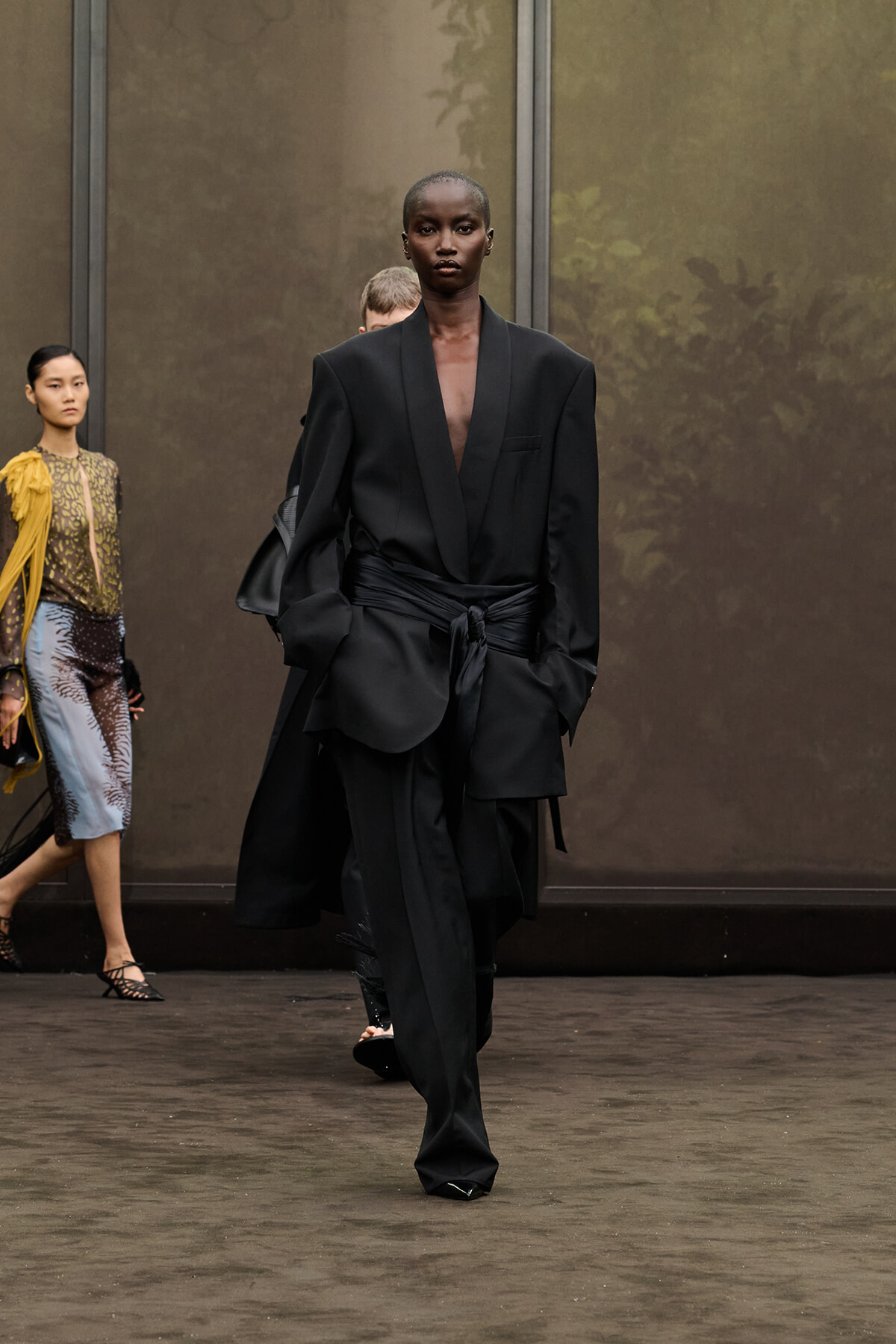 Model on the runway wearing an oversized black blazer with a wrapped waist and wide-leg pants, walking forward confidently.