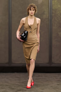 Model walking a runway in a tan sleeveless dress with buttoned placket, carrying a glossy black clutch and wearing red pointed heels