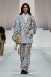 Model walks the runway in a light blue striped suit with brown floral embroidery, white shirt, and fluffy fur handbag accessory.