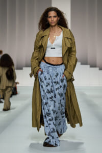 Runway model wearing a tan trench coat over a white crop top and blue striped floral pants.
