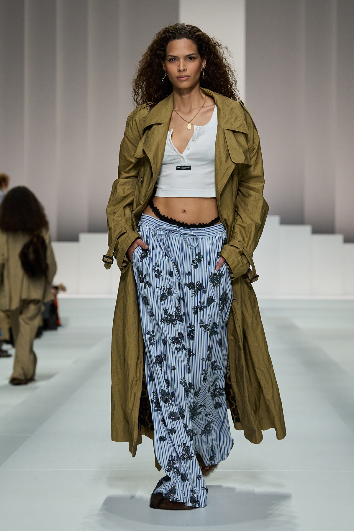 Runway model wearing a tan trench coat over a white crop top and blue striped floral pants.