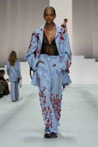 Model on runway wearing a light blue striped suit with red floral embroidery over a black lace top and gold necklace, walking forward confidently.