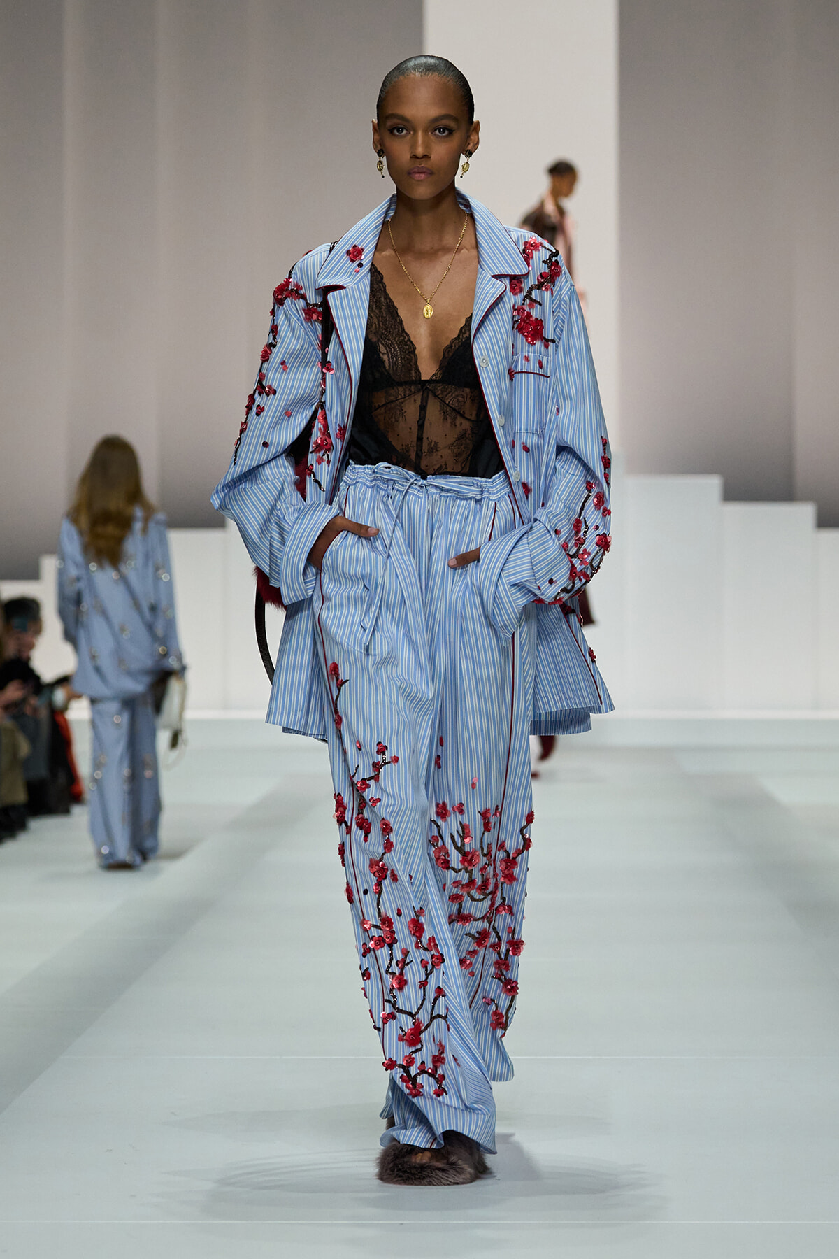 Model on runway wearing a light blue striped suit with red floral embroidery over a black lace top and gold necklace, walking forward confidently.