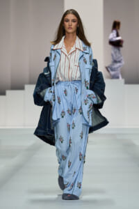 Model walking the runway in a light blue pinstriped suit with floral embroidery, denim jacket draped over her shoulders.