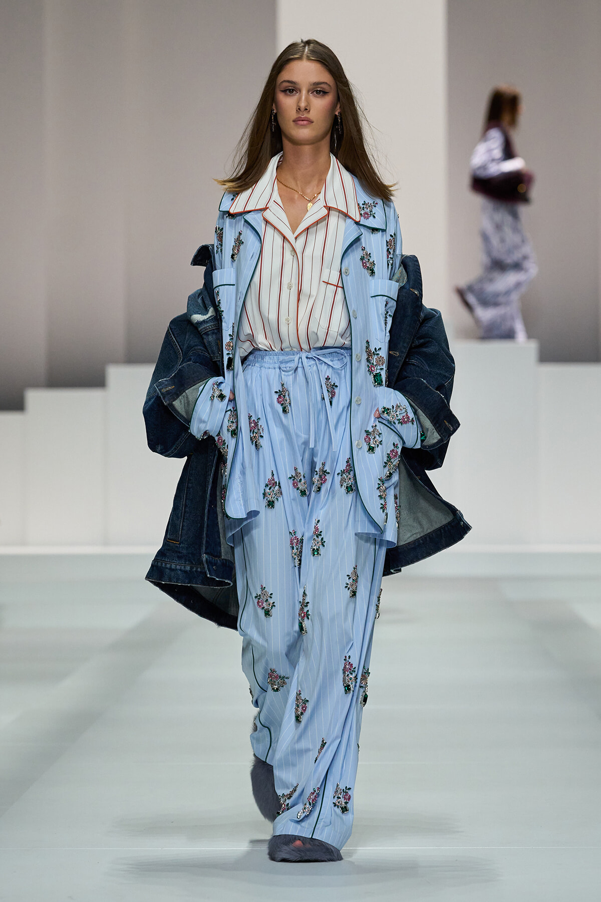 Model walking the runway in a light blue pinstriped suit with floral embroidery, denim jacket draped over her shoulders.