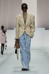 Model on a runway wearing a oversized beige pinstripe blazer with a sheer lace turtleneck and blue striped embroidered pants, sunglasses, and fur-handled handbag.