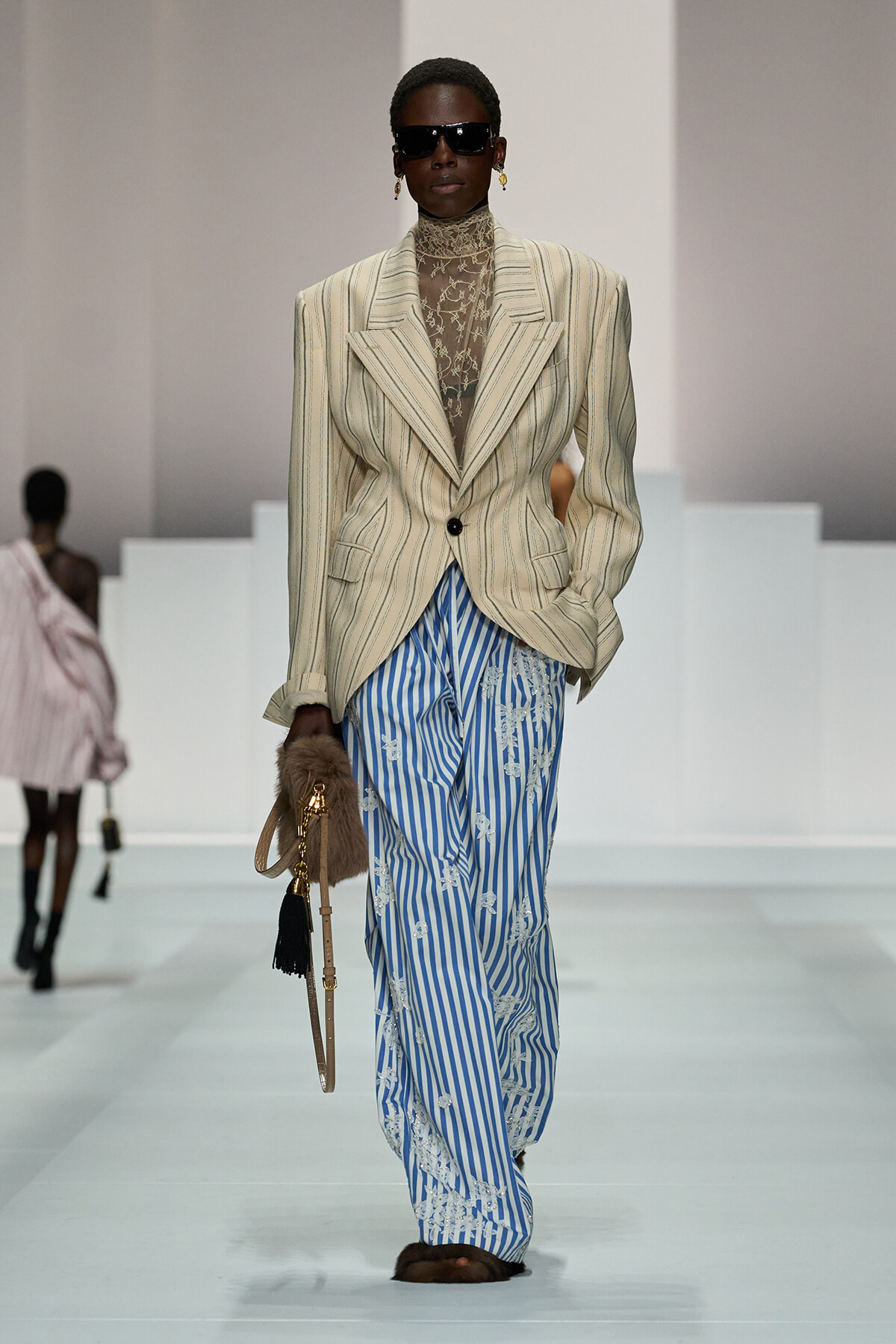 Model on a runway wearing a oversized beige pinstripe blazer with a sheer lace turtleneck and blue striped embroidered pants, sunglasses, and fur-handled handbag.