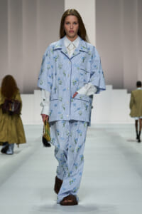 Model walks the runway in a light blue striped floral suit with a white shirt, carrying a yellow handbag with a black tassel.