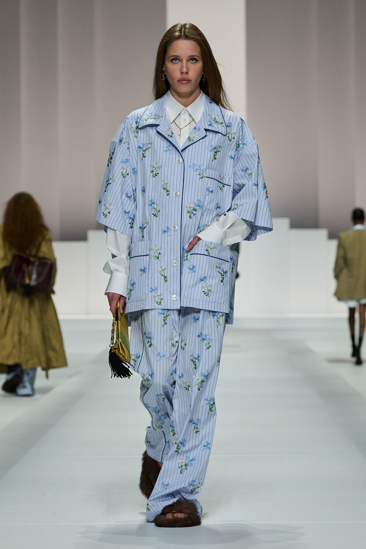Model walks the runway in a light blue striped floral suit with a white shirt, carrying a yellow handbag with a black tassel.