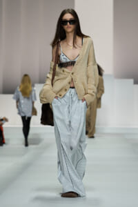 Model walking the runway in a chunky beige cardigan over a light blue lace-trim bralette and blue striped wide-leg pants, with dark sunglasses and a brown bag strap over the shoulder.