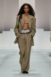 Model walking a runway in an oversized tan suit with a black bralette, high-waisted wide-leg pants, and a belt, carrying a shoulder bag.