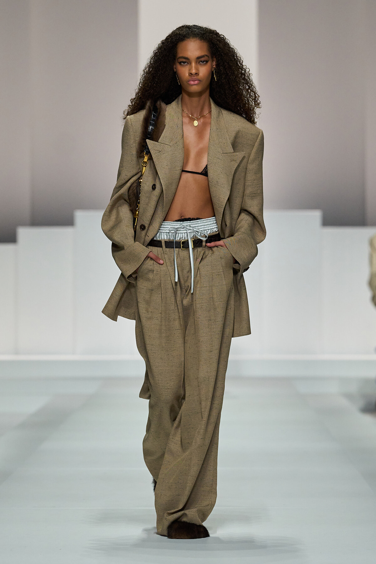 Model walking a runway in an oversized tan suit with a black bralette, high-waisted wide-leg pants, and a belt, carrying a shoulder bag.