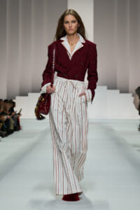 Model walking a fashion runway in a burgundy pinstripe double-breasted blazer with white pinstriped wide-leg pants, carrying a burgundy handbag.