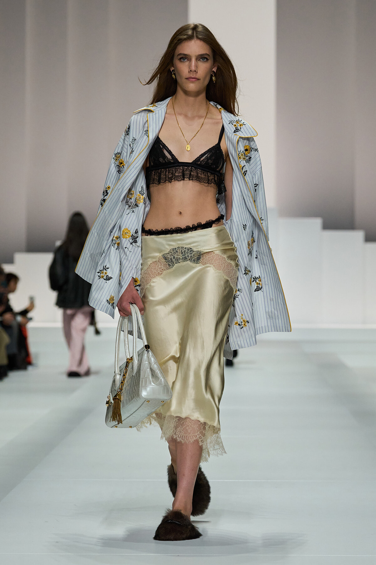 Model walks the runway wearing a black lace bralette with a champagne satin skirt that has a lace hem, a blue striped embroidered jacket draped over shoulders, and a silver handbag.