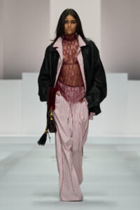 Female model walks a runway in pink striped wide-leg pants, burgundy lace top under a black leather jacket, holding a small tassel handbag.