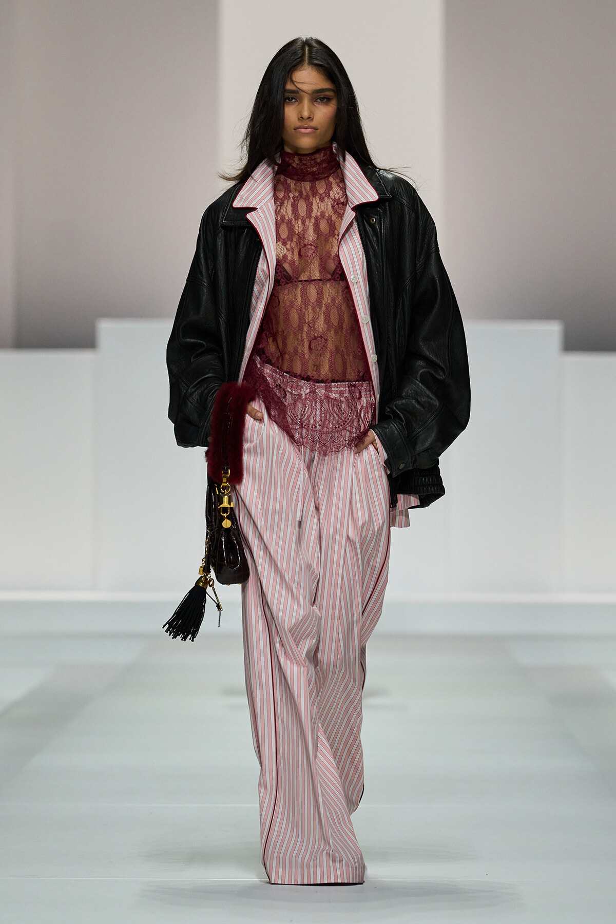 Female model walks a runway in pink striped wide-leg pants, burgundy lace top under a black leather jacket, holding a small tassel handbag.