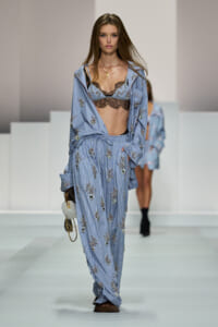 Model walking a fashion show in a light blue striped, embroidered pajama-style suit with a black lace bralette showing beneath.