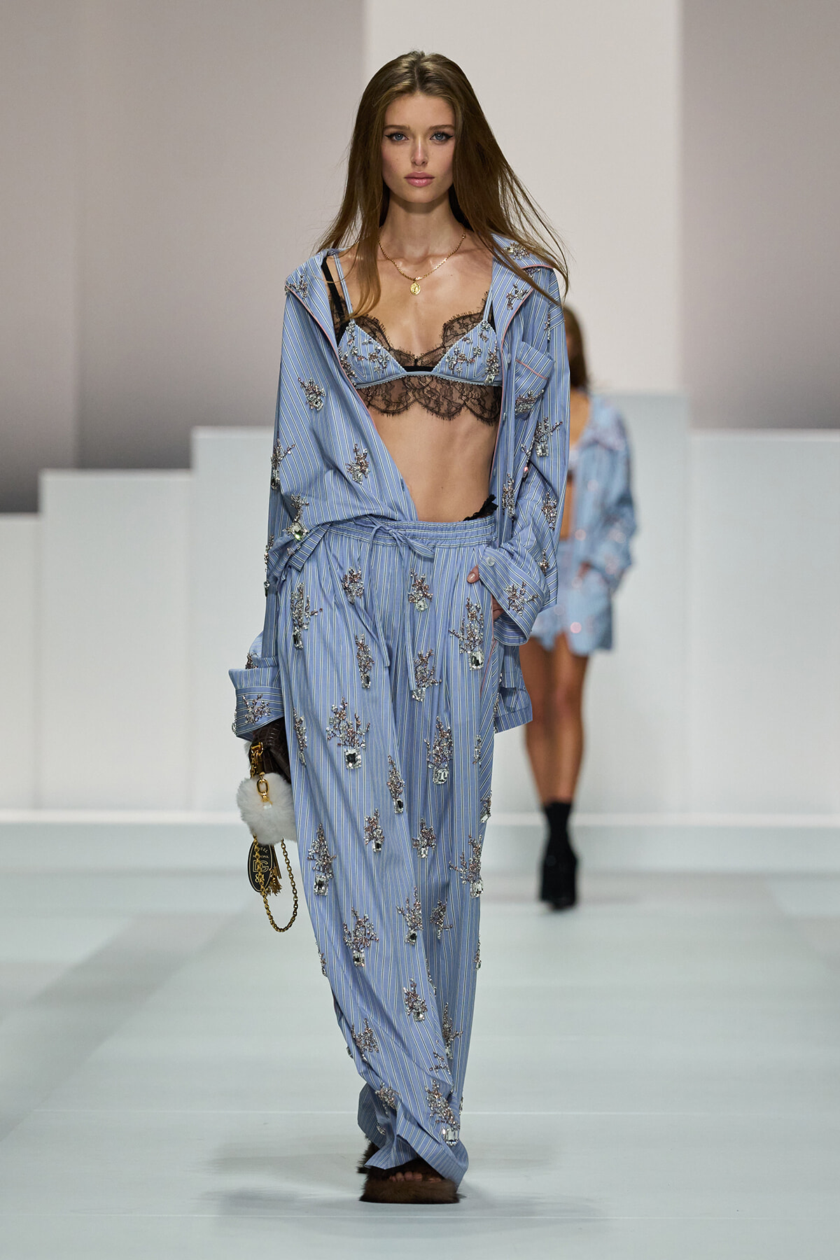 Model walking a fashion show in a light blue striped, embroidered pajama-style suit with a black lace bralette showing beneath.