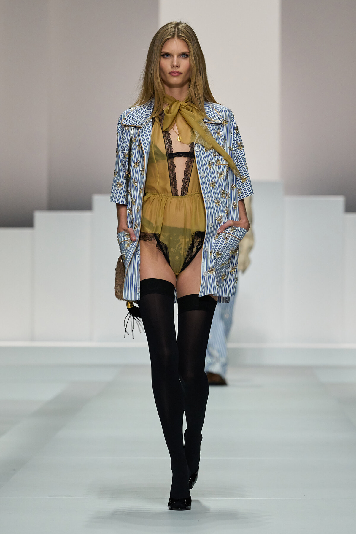 Model on runway wearing sheer mustard lingerie with black lace, a blue striped embroidered coat, and black thigh-high stockings.