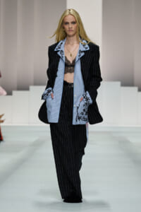 Model walks a runway wearing a black pinstripe suit layered over a light blue striped shirt with embroidered cuffs; black lace bralette visible beneath.