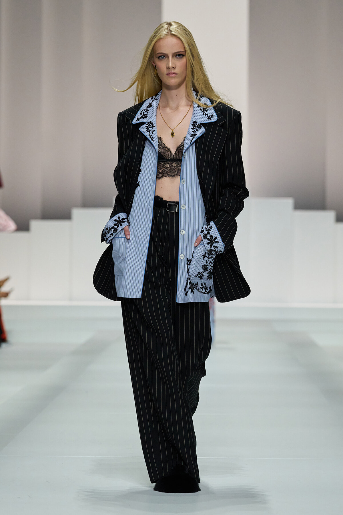Model walks a runway wearing a black pinstripe suit layered over a light blue striped shirt with embroidered cuffs; black lace bralette visible beneath.