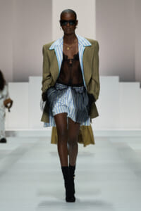 Runway model in an olive pinstripe blazer over a blue striped shirt, sheer black lace top, and striped shorts with black mesh overlay.