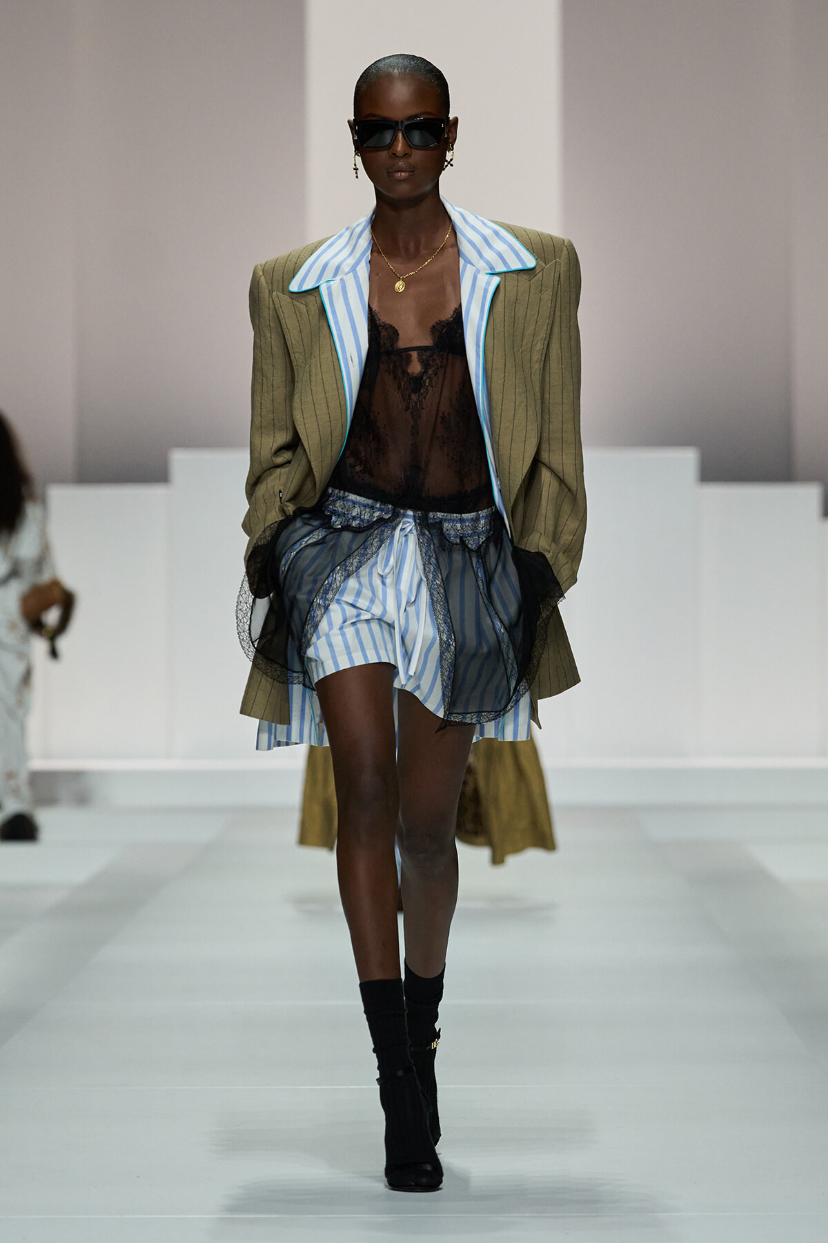 Runway model in an olive pinstripe blazer over a blue striped shirt, sheer black lace top, and striped shorts with black mesh overlay.