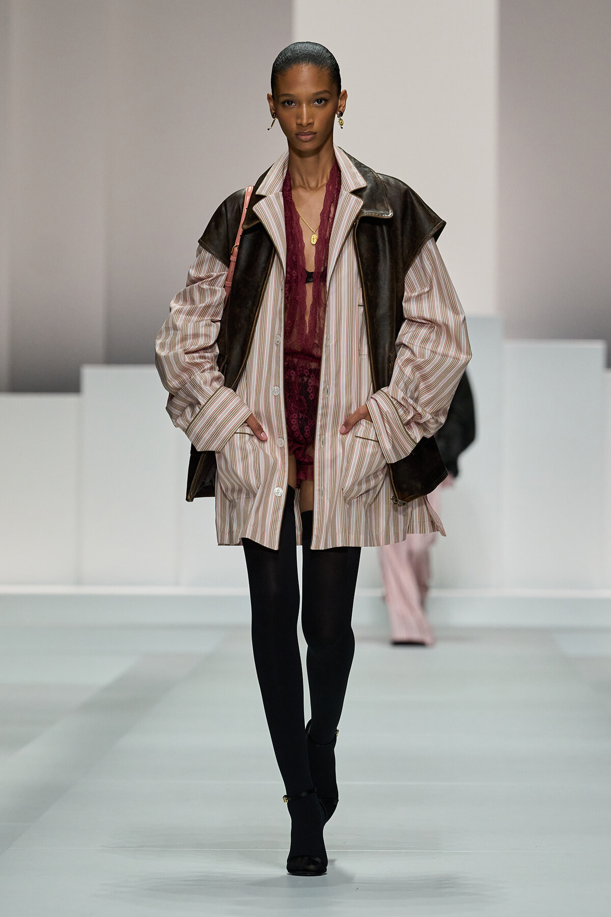 Female model walks the runway wearing a striped oversized coat over a burgundy lace dress, black tights and ankle boots.