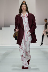 Runway model wears a burgundy blazer over a white striped pajama-style suit with red floral embroidery, walking the catwalk.