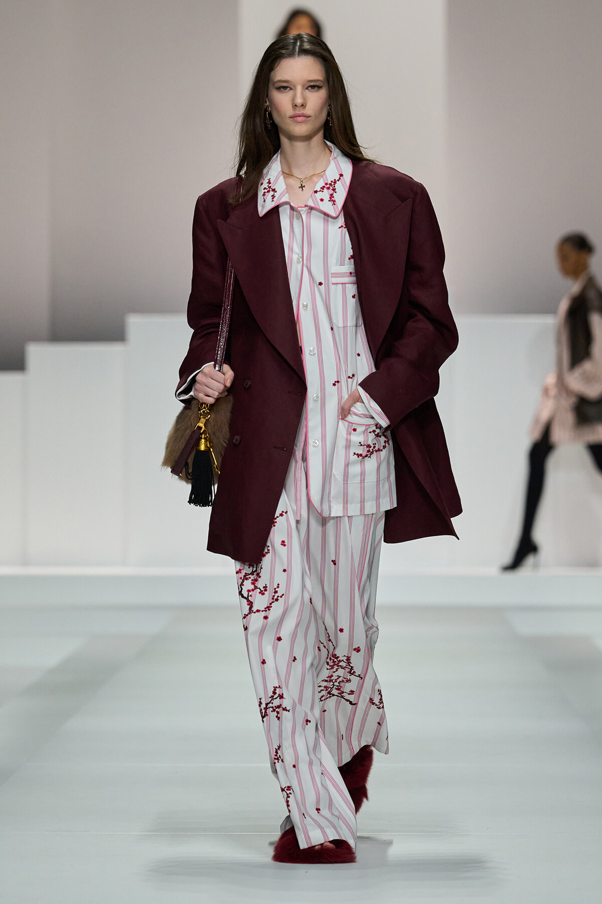 Runway model wears a burgundy blazer over a white striped pajama-style suit with red floral embroidery, walking the catwalk.