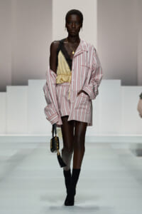 Runway model in an oversized pink striped shirt worn as a dress, with a yellow satin top and black lace, carrying a small black handbag with tassel.