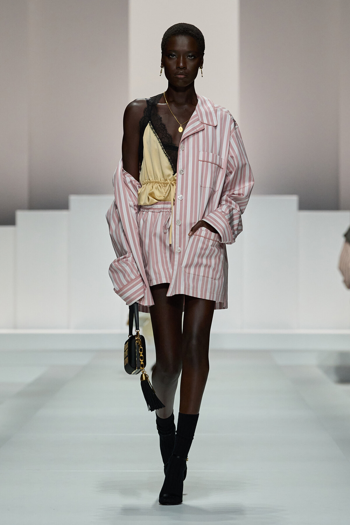 Runway model in an oversized pink striped shirt worn as a dress, with a yellow satin top and black lace, carrying a small black handbag with tassel.